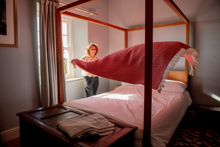 A woman makes the bed in a clean, sunlit bedroom with a four-poster bed and wooden chest at the foot.
