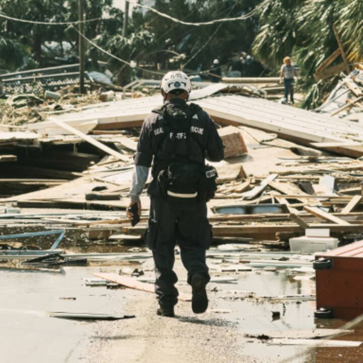 A rescue worker walks through debris in a disaster-stricken area, with fallen structures and scattered wreckage.