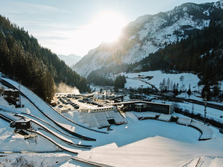 Snowy ski resort with ski jumps and an empty stadium, surrounded by mountains and forest, under a bright sunrise.