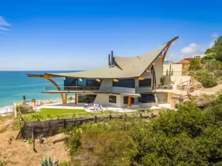 A modern, uniquely designed beachfront house with a large angular roof and glass walls overlooking the ocean.