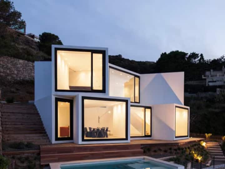 Modern, multi-level white house with large, illuminated windows and an outdoor swimming pool at dusk.