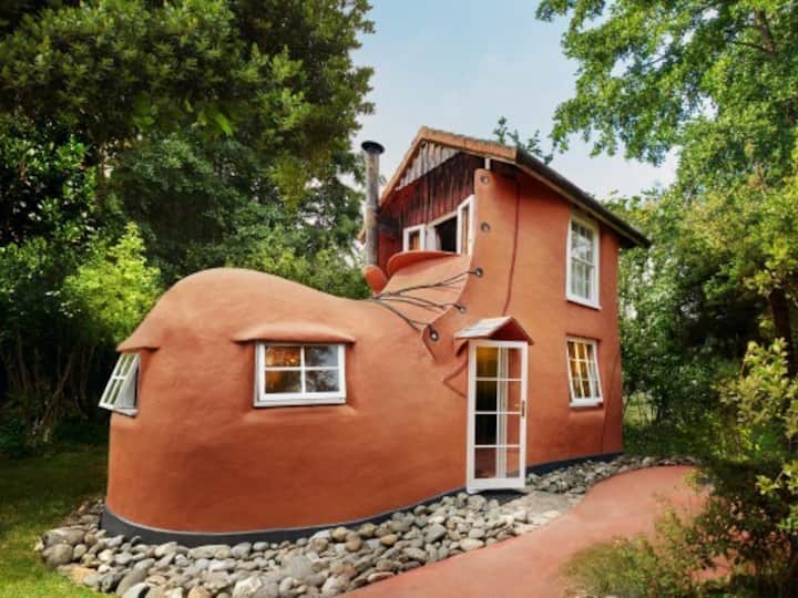 A unique house designed to look like a shoe, with a red exterior, multiple windows, and a white door, surrounded by lush greenery.
