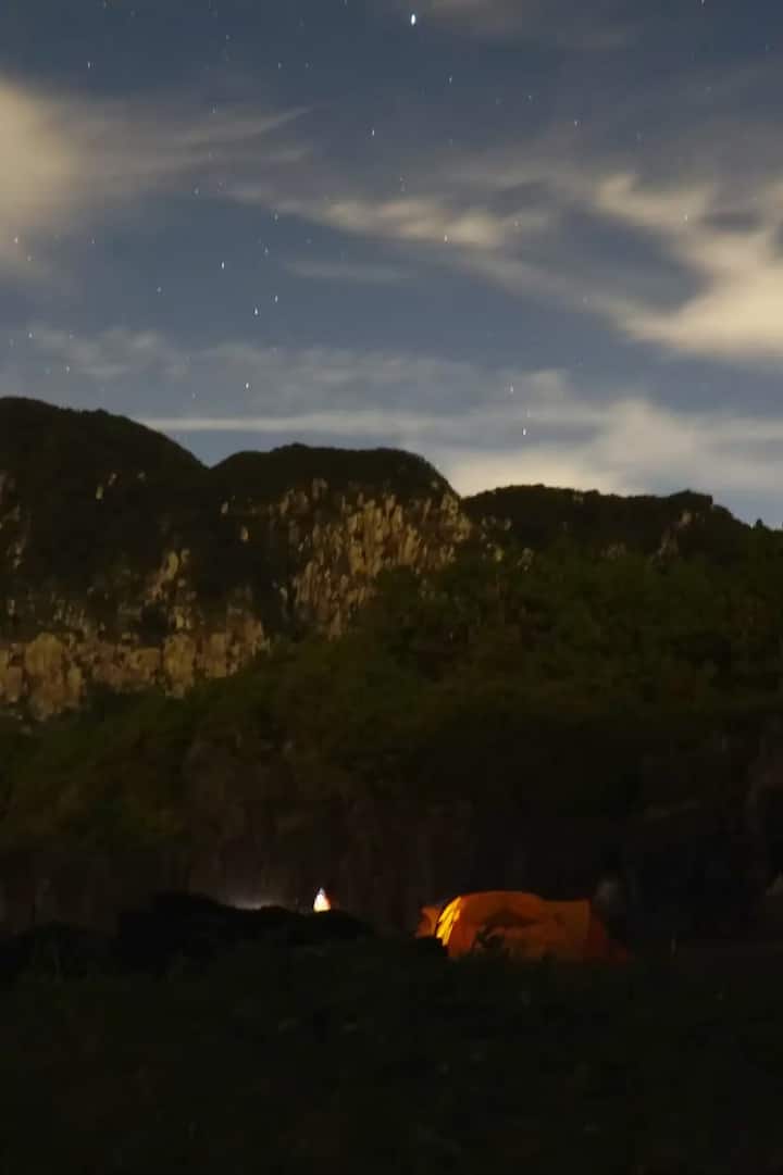 Starry night sky above a mountain range with a lit tent at the base.