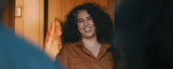 A woman with dark curly hair wearing a brown shirt smiles from a doorway with a wooden wall in the background