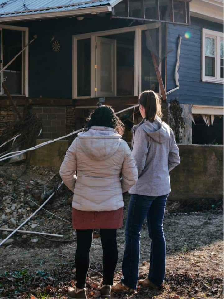 Two women, both with dark hair, have winter jackets on and are looking at a damaged home from a recent hurricane.