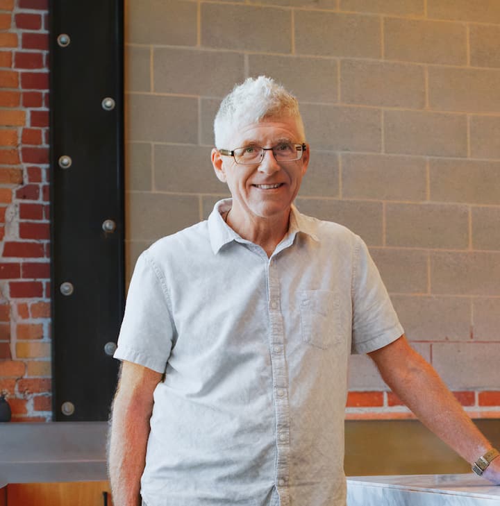 A Host standing in a modern industrial kitchen with brick and cement walls.