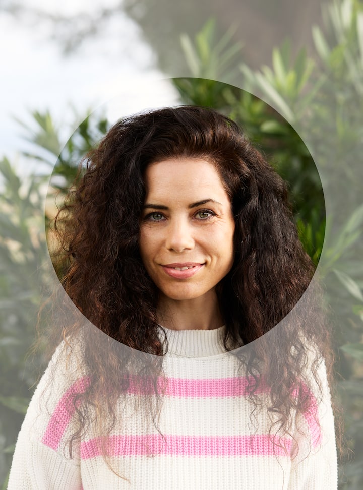 Close-up portrait of a person with dark curly hair, dressed in a white sweater with pink stripes, with greenery in the background.