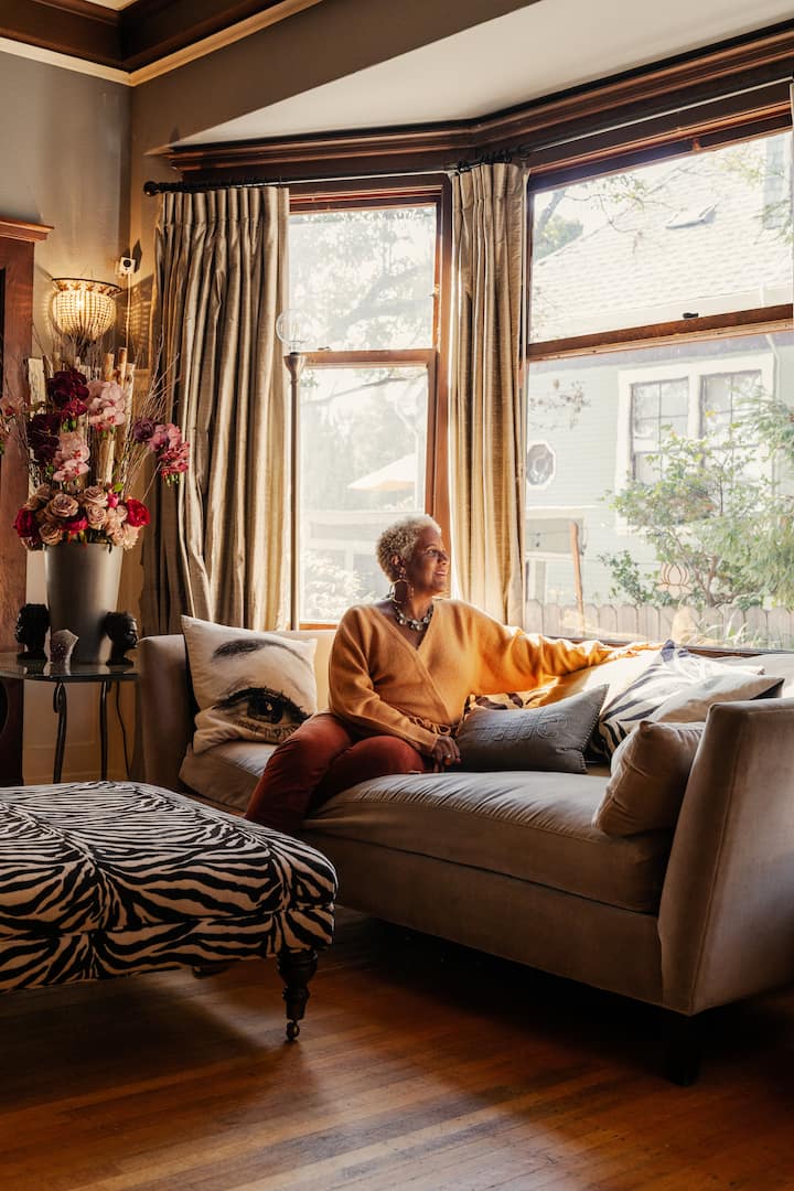 Woman sitting on beige suede couch looks out large bay window.