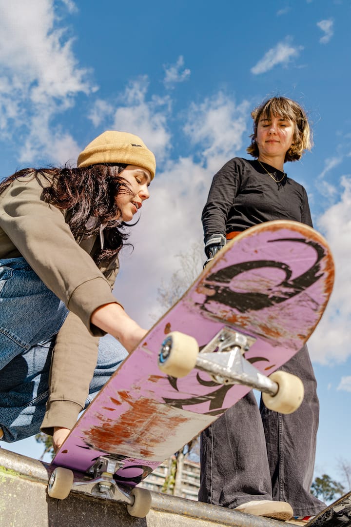 Close-up of a skateboarder in a yellow beanie getting ready to ride a pink skateboard, with another person standing next to them.