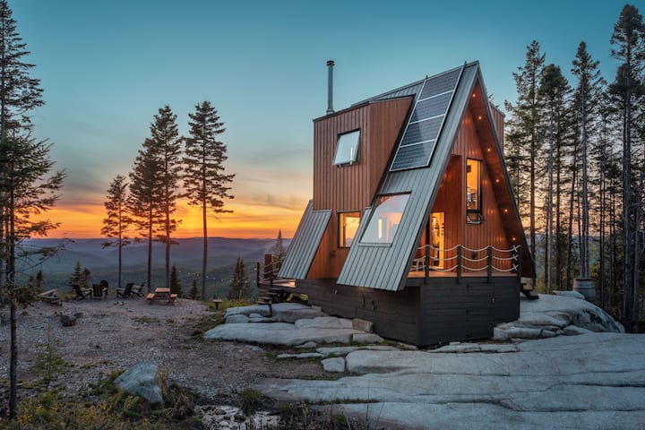 Cabin sits atop stone cliff overlooking mountains at sunset