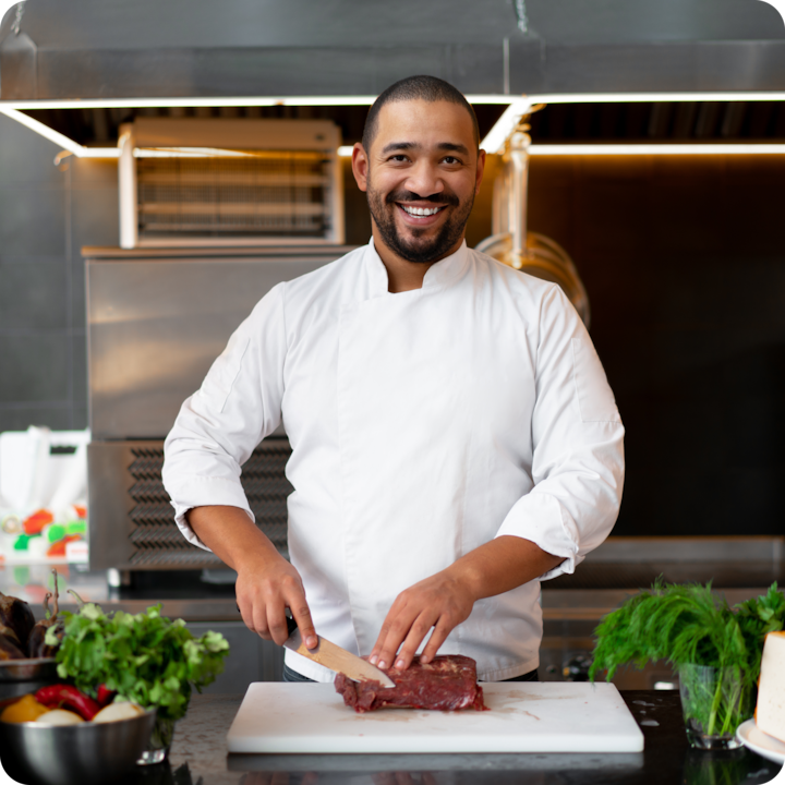 Chef in a white uniform slicing a piece of raw meat on a cutting board in a professional kitchen.