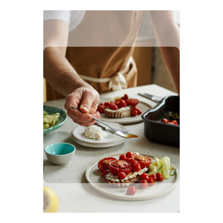 Person in a brown apron preparing toast with tomatoes and a dollop of cheese on a kitchen counter.