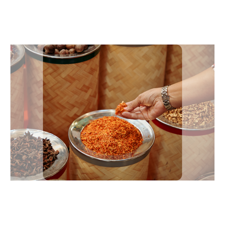 A hand picking up dried orange herbs from a silver bowl, surrounded by baskets full of other spices.