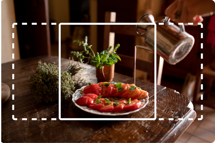 A beautifully presented dish of tomato slices with herbs on a white plate, placed on a rustic wooden table alongside a small potted plant and dried herbs, with a hand pouring water from a kettle in the background.