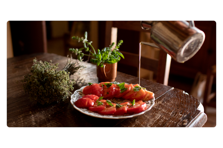 Close-up of a plate with tomato slices topped with herbs, set on a rustic wooden table with a pot of fresh green herbs and a metal kettle pouring liquid.