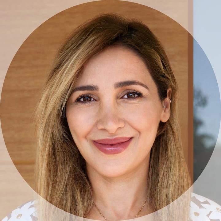 A professional profile photo showing a person with long blonde hair, wearing a white top with floral patterns, standing in front of a wooden and glass door background.