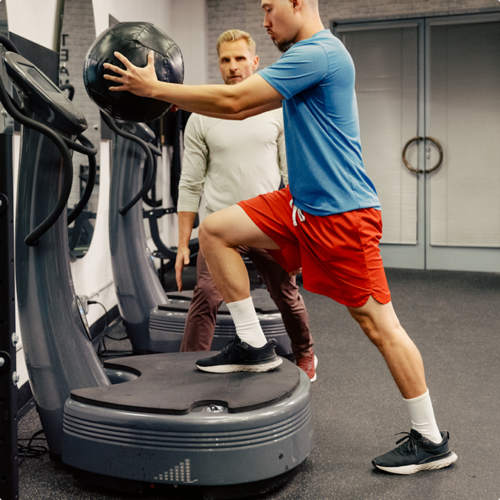 A man in red shorts and blue shirt holds a medicine ball on a vibration plate machine while a host supervises in gym.