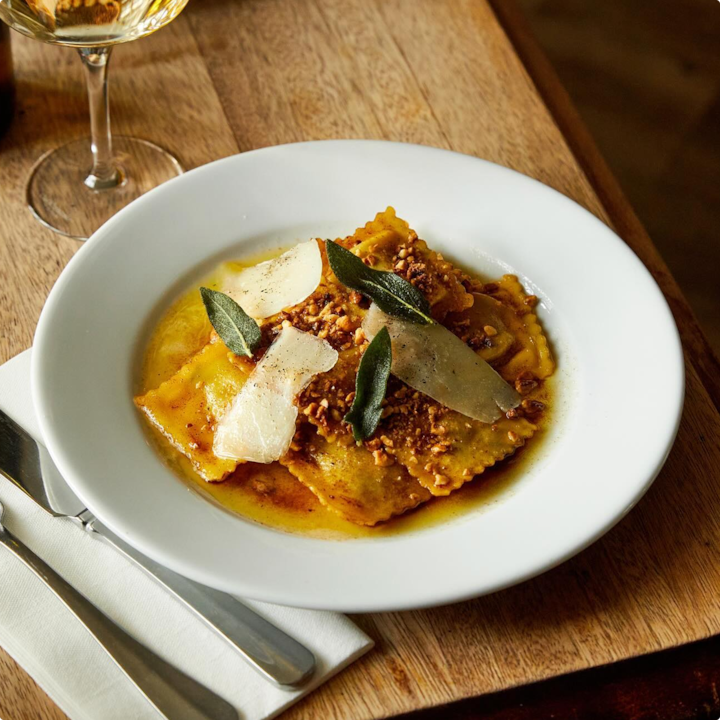 A dish of ravioli topped with cheese shavings and sage leaves, served with a glass of white wine.