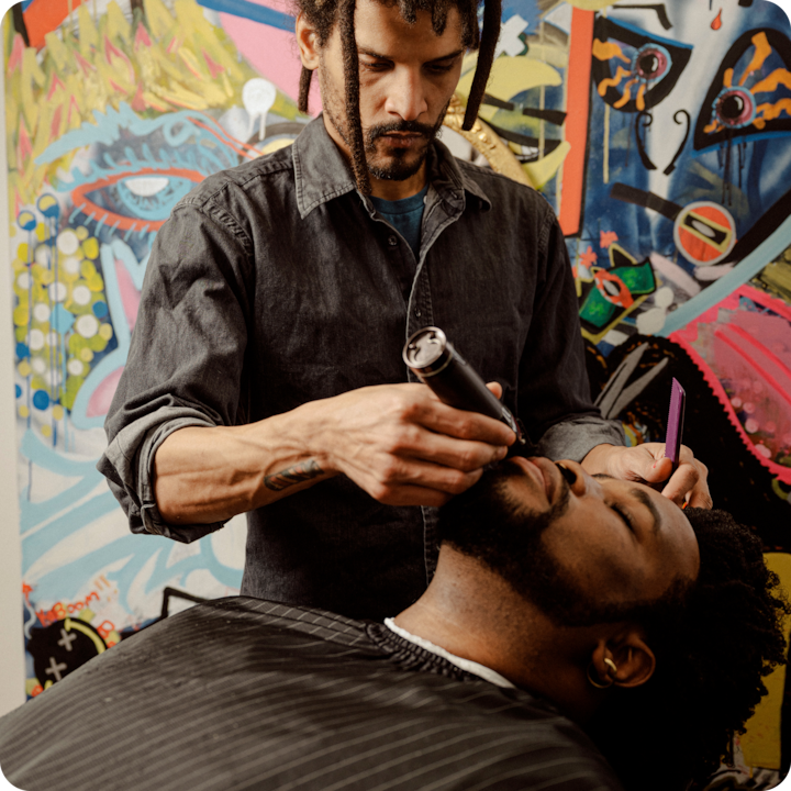 Close-up of a barber trimming a client's beard, with a detailed graffiti-style mural on the wall behind them.