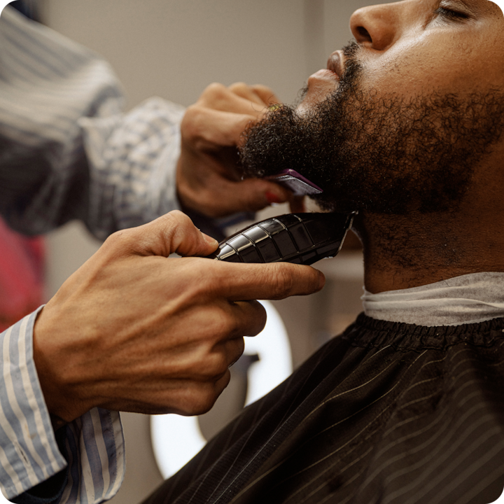 Close-up of a person trimming another person's beard with an electric razor in a barber shop.