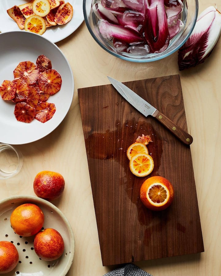 Top-down view of a kitchen setup with a cutting board and knife, freshly sliced blood oranges, and bowls of endive and blood orange slices.