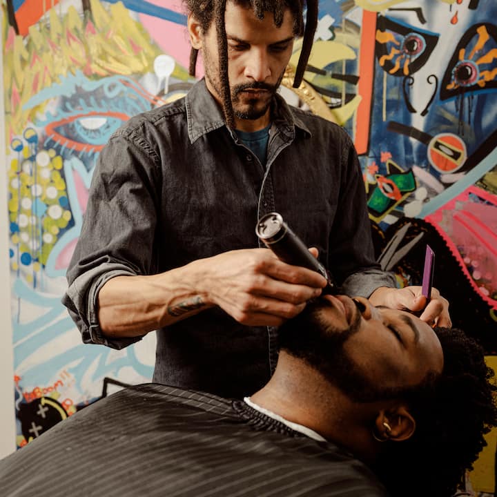 Un homme se fait tailler la barbe par un barbier devant des graffitis colorés, soulignant l'importance de la photographie professionnelle pour les offres Airbnb.