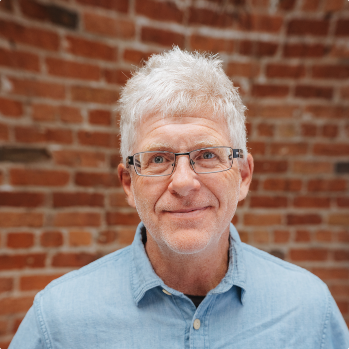 Plan rapproché d'une personne aux cheveux blancs et vêtue de façon décontractée. Un mur en brique se trouve à l'arrière-plan.