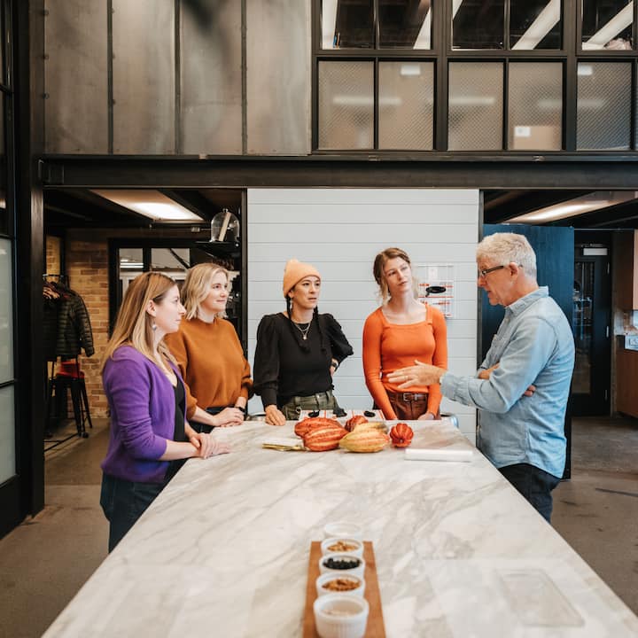Cinq personnes sont debout autour d'une table en marbre sur laquelle sont posés des cabosses de cacao et des plats de condiments. Elles discutent dans un espace de travail contemporain.