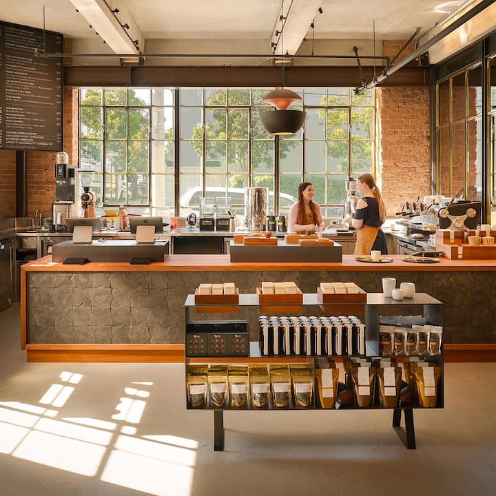 Deux femmes discutent derrière le bar d'un café moderne. De grandes fenêtres se trouvent à l'arrière-plan. Divers produits liés au café sont exposés sur le comptoir.