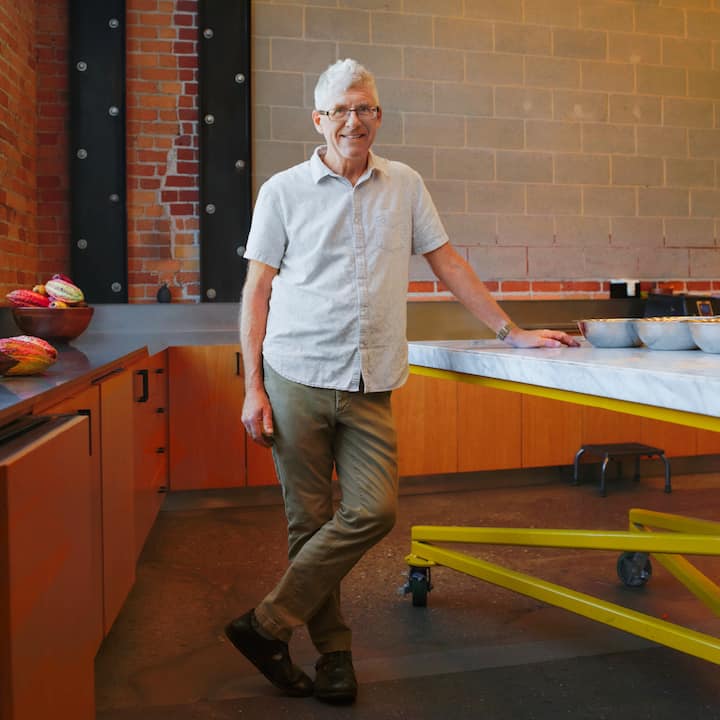 Un homme porte une tenue décontractée : une chemise légère et un pantalon kaki. Il se tient à côté d'un long plan de travail dans une cuisine moderne, montrant un espace dédié à la préparation de spécialités culinaires.