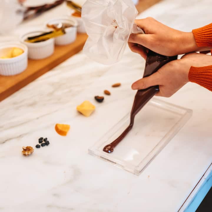 Plan rapproché d'une personne qui remplit un moule de chocolat. Des ingrédients, comme des fruits à coque et des fruits secs, se trouvent tout autour sur un plan de travail en marbre.