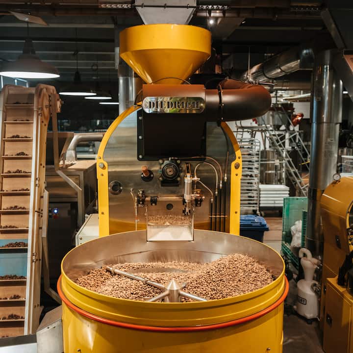 Une grande machine jaune servant à torréfier le café se trouve dans une usine. Elle est remplie de grains de café cru.