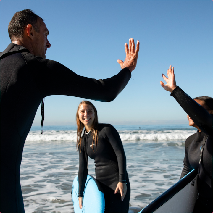 Surf instructor high-fives two students on the beach with their surfboards after a successful session.