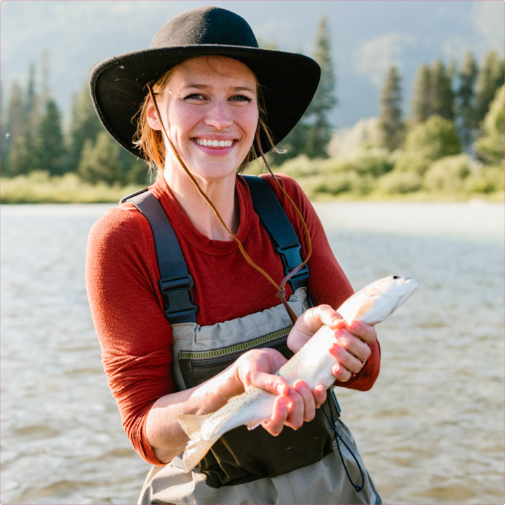 Person in red shirt and black hat holding a fish while standing in a lake with trees and mountains in the background.