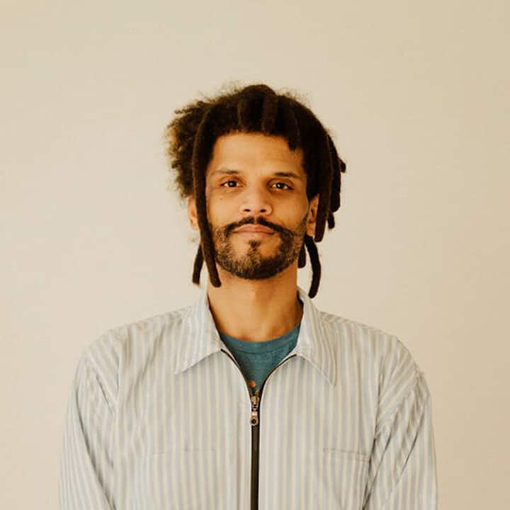 Un homme portant une chemise rayée bleu clair avec des motifs verts et bleus prend la pose devant un arrière-plan épuré.