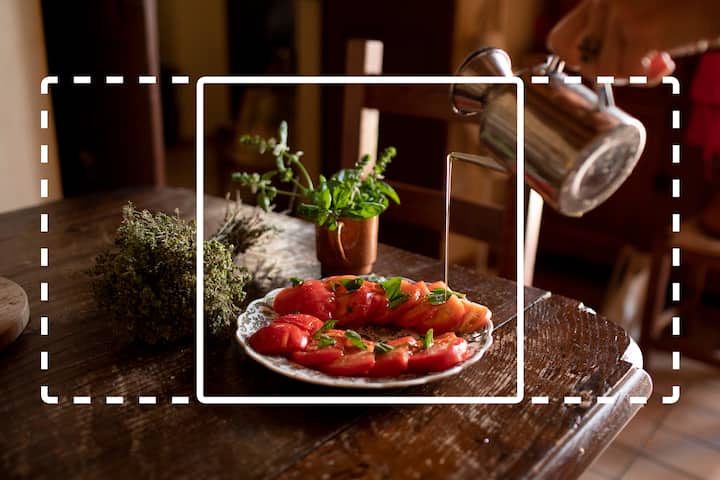Une assiette blanche, qui contient un plat de tranches de tomates aux herbes, est joliment présentée sur une table rustique en bois. À côté se trouvent une petite plante en pot et des herbes séchées. Une main verse l'eau d'une bouilloire en arrière-plan.