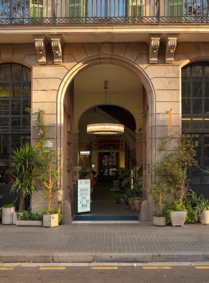 Photo montrant l'extérieur avant une expérience Airbnb, avec l'entrée d'un bâtiment dotée d'une arche. Des plantes et une pancarte indiquant les options de petit déjeuner sont présentes.