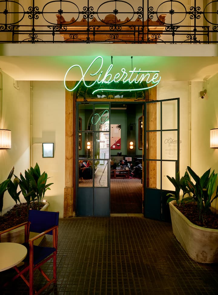 Entrée d'un lieu tendance avec une enseigne lumineuse indiquant « Libertine ». L'espace intérieur est orné de plantes et doté de sièges.