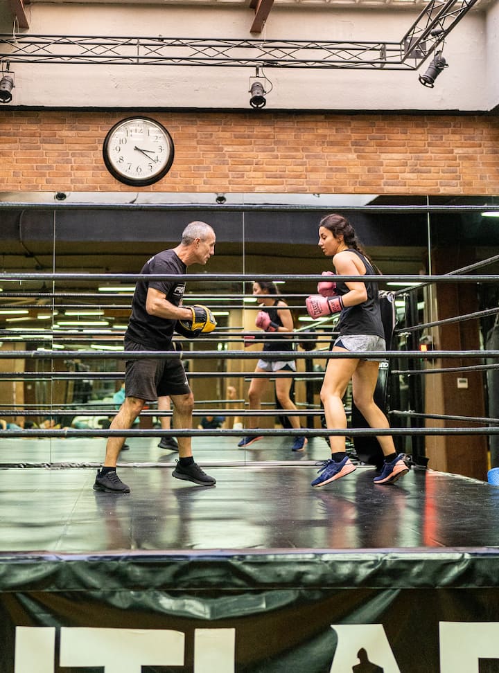 A boxing coach instructs a woman in the ring, both wearing gloves, with another woman visible in the background practicing boxing.