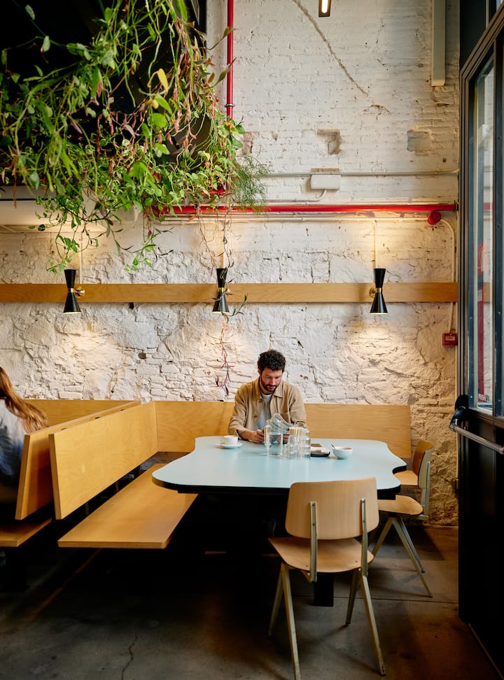 A cozy cafe with a rustic white brick wall, a wooden bench, a light blue table, and hanging plants above. A person is seated alone, working with a cup of coffee and a water jug on the table.