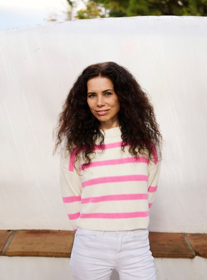 Host wearing a white sweater with pink stripes and white pants, standing outdoors in front of a light backdrop with greenery visible in the background.