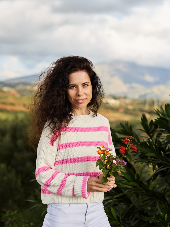 A person standing outdoors holding a small bouquet of colorful flowers, wearing a white sweater with pink stripes and white pants, with lush greenery and a mountain landscape in the background.