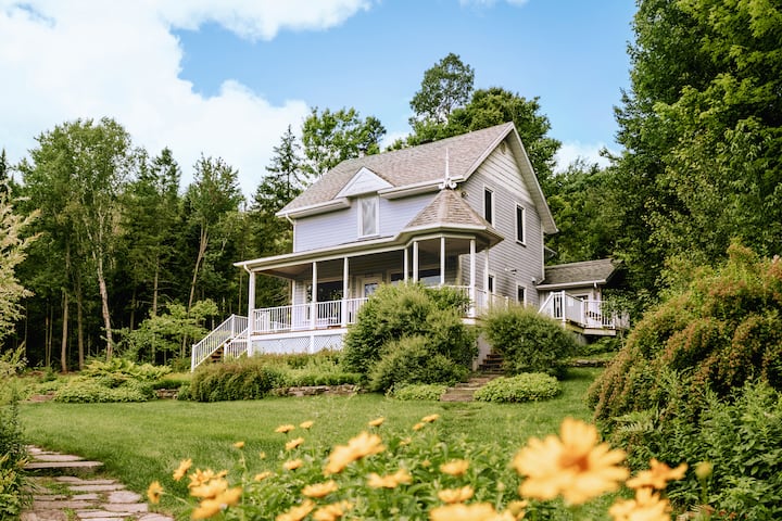 A house sits alone near a forest, surrounded by grassy fields and tall trees on a sunny day.