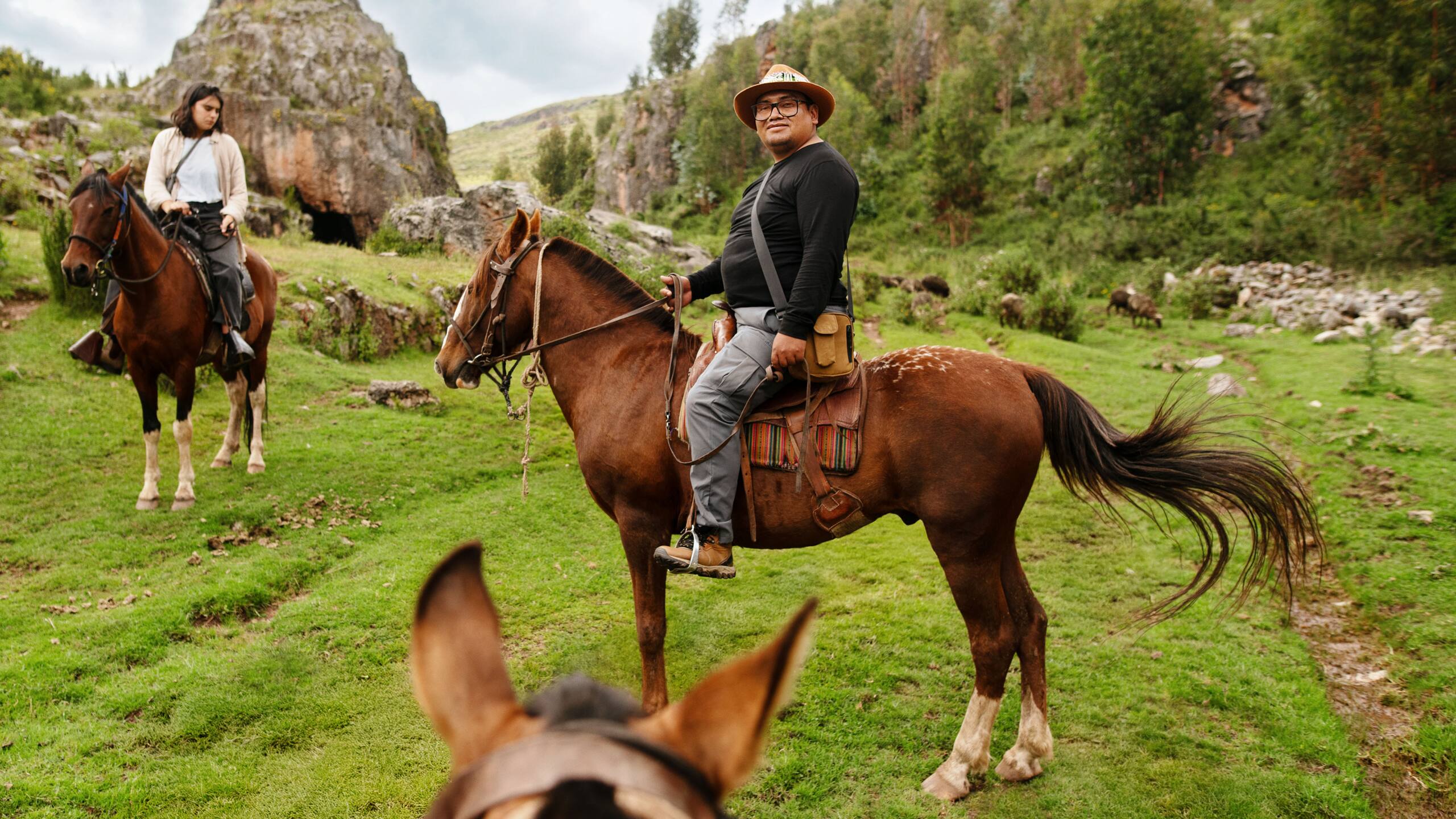 Dos personas montan a caballo en un paisaje montañoso verde, con formaciones rocosas al fondo y un grupo de animales pastando a lo lejos.