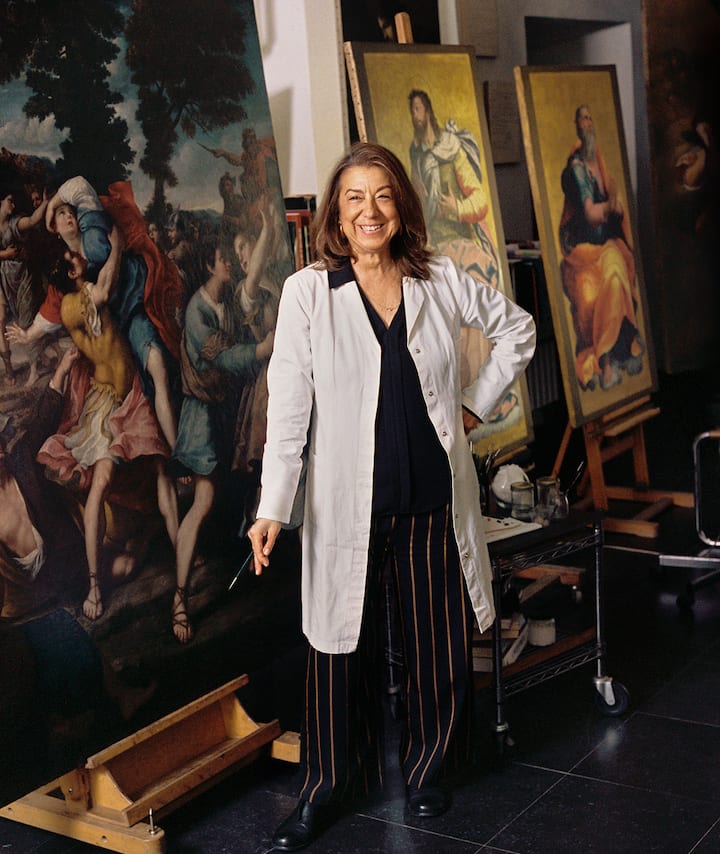 An art restorer in a white coat stands proudly in front of framed art pieces.