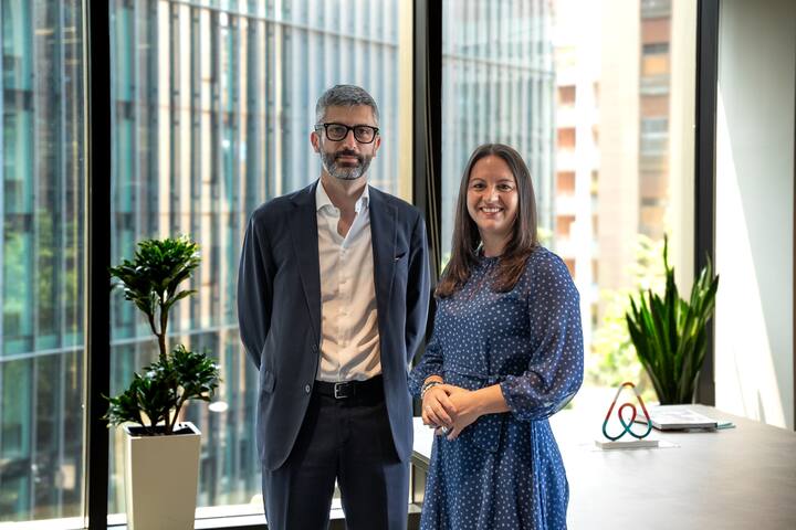 Two people standing in a bright office space with large glass windows, showcasing modern buildings and greenery outside. A desk in the foreground holds a colorful Airbnb logo sculpture.