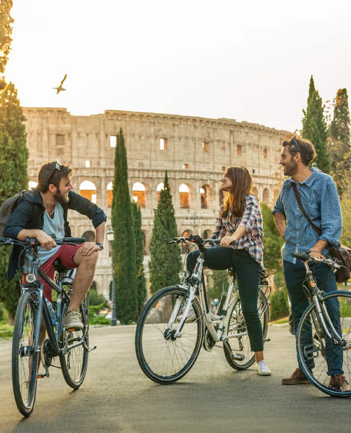 Three people on bicycles pause in front of the Colosseum in Rome at sunset, with tall cypress trees framing the scene.