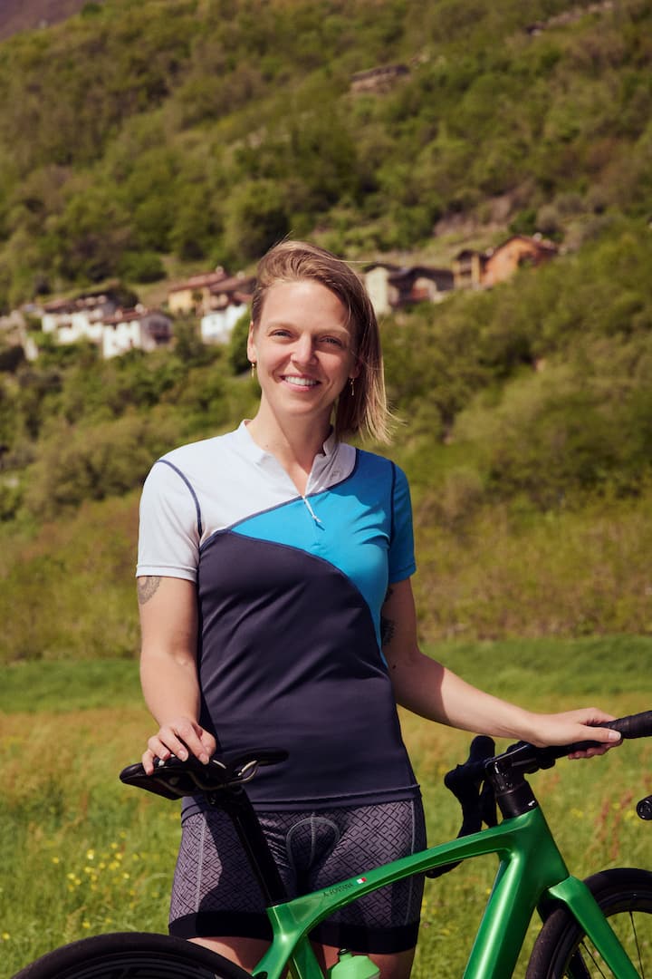 A person wearing a blue and black cycling jersey stands next to a green bicycle in a lush, rural landscape with hills and small houses in the background.