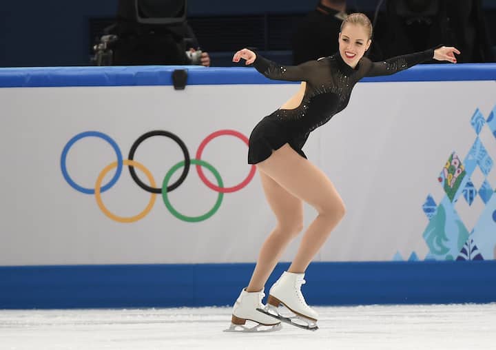 Figure skater in a black costume gliding on ice in front of the Olympic rings and event signage.