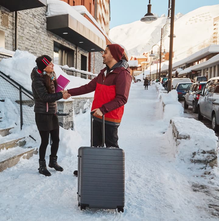A traveler in a red winter jacket with a suitcase shakes hands with a host outside a snowy chalet-style building in a mountain resort.